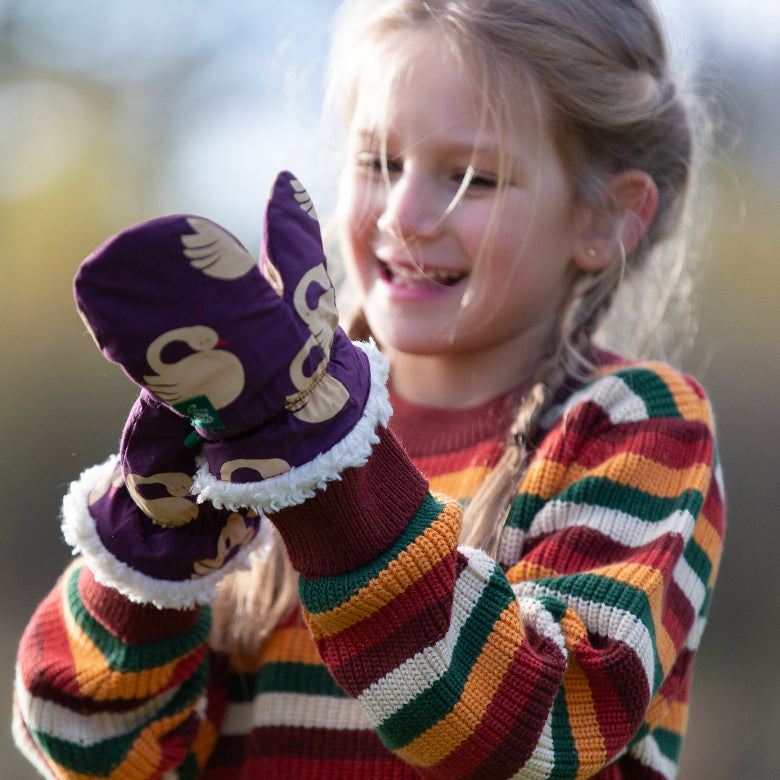 A young girl outdoors wearing a colorful striped knit sweater and purple winter mittens with a cartoon duck print and white faux-fur trim, holding something in her hands.