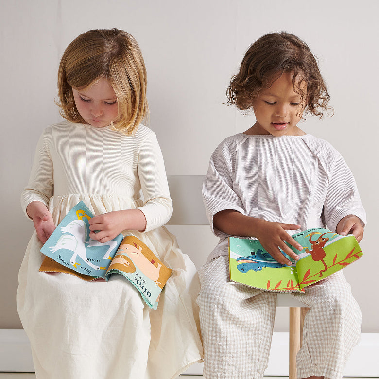Two young children sitting side by side on a white bench, each holding an open colorful children's book. The child on the left wears a cream long-sleeve dress, and the child on the right wears a white waffle-knit top and light checkered pants.