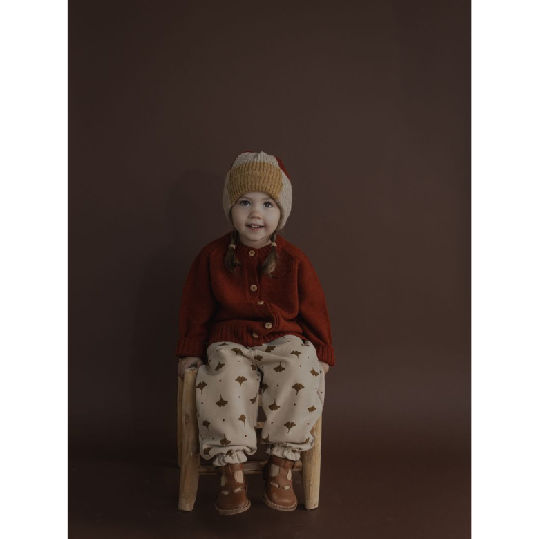 A young child sitting on a small wooden stool against a dark brown studio background, wearing a rust red knit cardigan, cream knit pants with a brown pattern, and a beige knit beanie.
