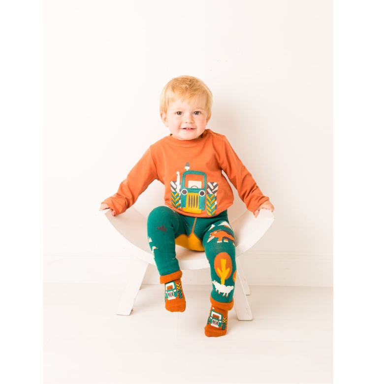 Child wearing an orange shirt with a tractor design and green pants with animal prints, sitting on a white chair.