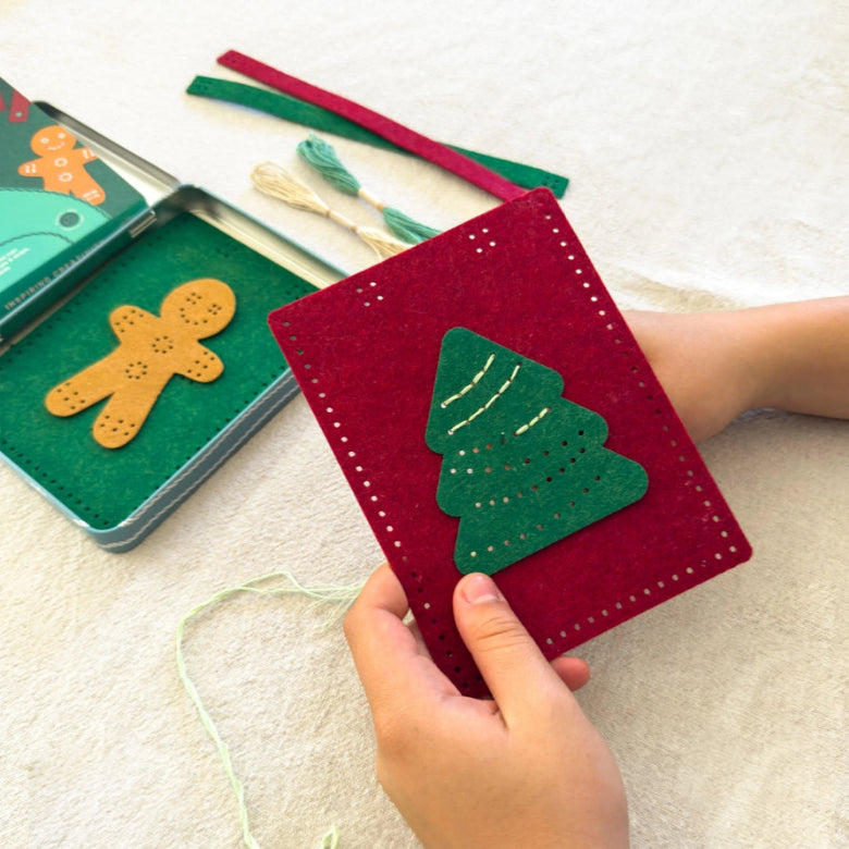 A child's hands hold a red felt pocket notebook with a green felt Christmas tree applique. In the background, a felt craft kit with a gingerbread man piece and ribbon bookmarks lies on a light beige fabric surface.