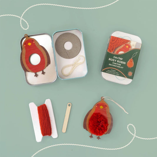 A flat-lay view of a 'Rosy Robin' felt decoration kit on a sage green background. The kit includes a felt robin ornament, a metal tin with felt and string, a felt spinner, a spool of red yarn, a wooden stick, and the product packaging.
