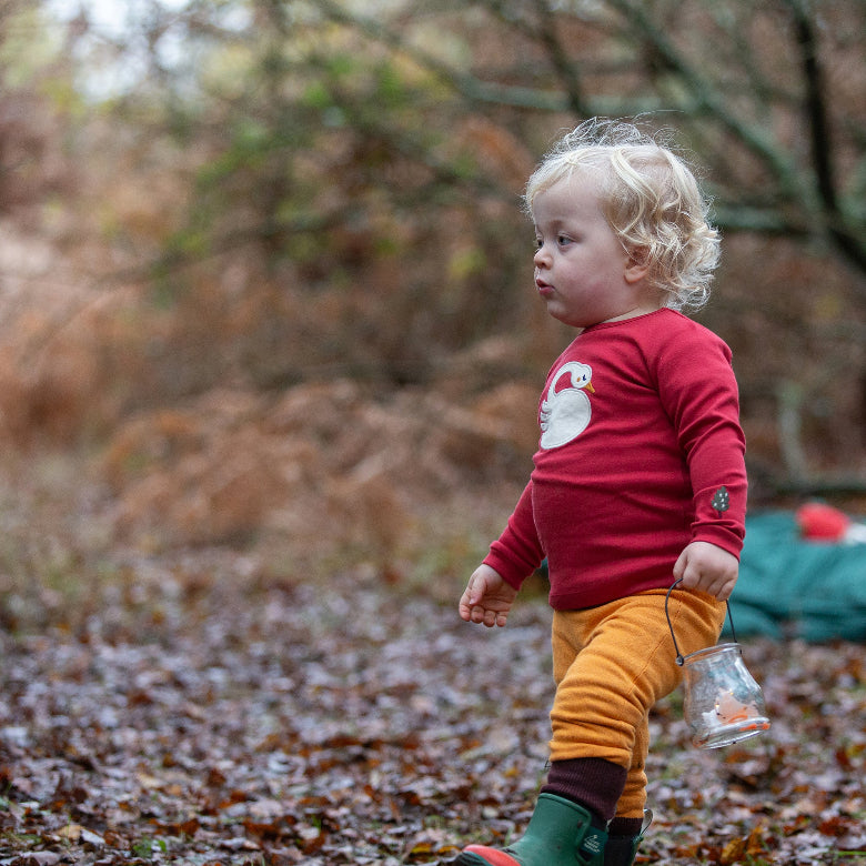 Child in a red shirt and yellow pants walking outdoors on a leaf-covered ground.