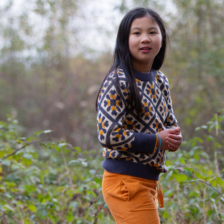A young girl with long dark hair stands outdoors in a wooded setting, wearing a navy, cream, and mustard geometric patterned knit sweater and orange pants.