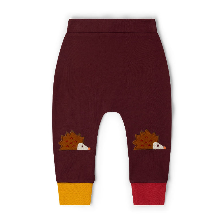 A pair of dark burgundy baby pants with two embroidered hedgehogs on the lower legs and contrasting yellow and red ankle cuffs, shown flat on a white background.