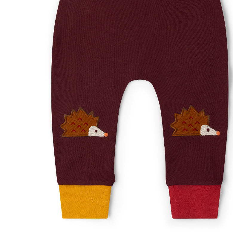 A close-up of a pair of burgundy baby pants featuring embroidered hedgehogs on each leg and contrasting yellow and red cuffs, shown against a plain white background.