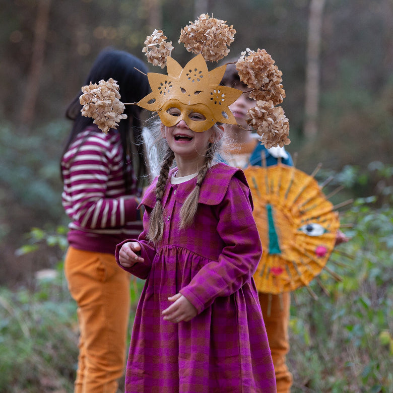 A child wearing a purple dress and a gold mask with dried flower decorations stands outdoors in a wooded setting, with other children partially visible in the background.