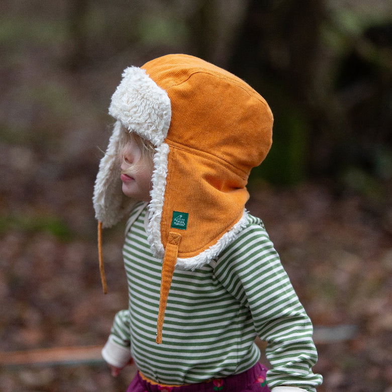 A toddler wearing an orange corduroy aviator-style winter hat with white faux-fur trim, photographed outdoors in a blurred forest setting.