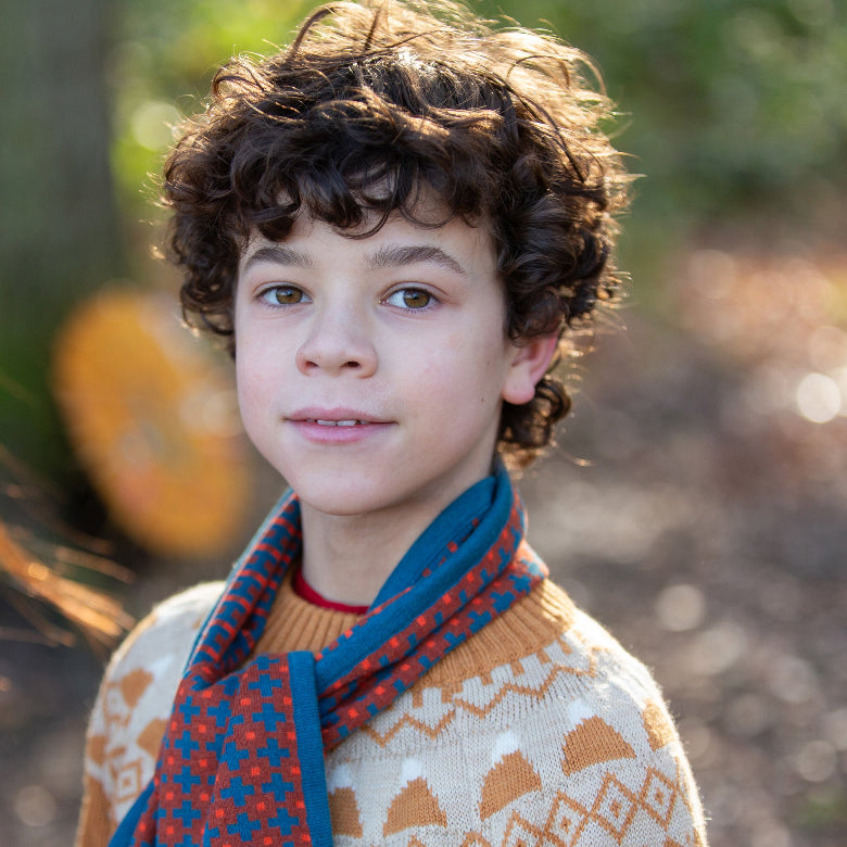 A close-up portrait of a young boy with curly brown hair wearing a cream knit sweater with orange geometric patterns and a blue scarf with red and orange accents, set against a blurred outdoor background.