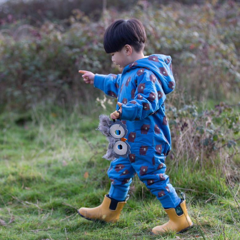 A young child wearing a blue hooded one-piece jumpsuit with a brown bear pattern and yellow rain boots, standing in a grassy outdoor setting and pointing to the left while holding a gray plush toy.
