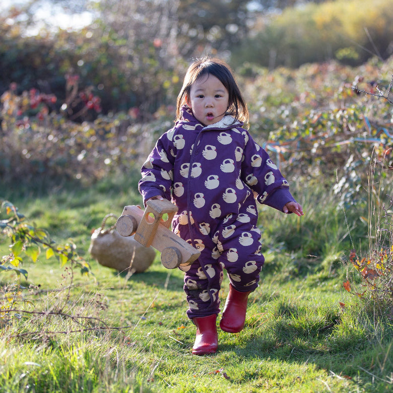 A toddler girl wearing a purple hooded one-piece snowsuit with a white monkey print is running outdoors on a grassy hill while holding a wooden toy truck.