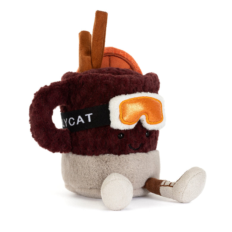 A plush toy shaped like a hot cocoa mug with a smiling face, wearing orange goggles and brown boots, on a solid white background.