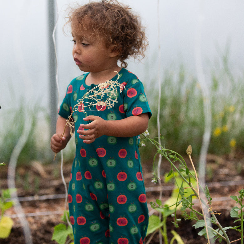 Child in a garden wearing a green dress with red and pink patterns, holding a plant.