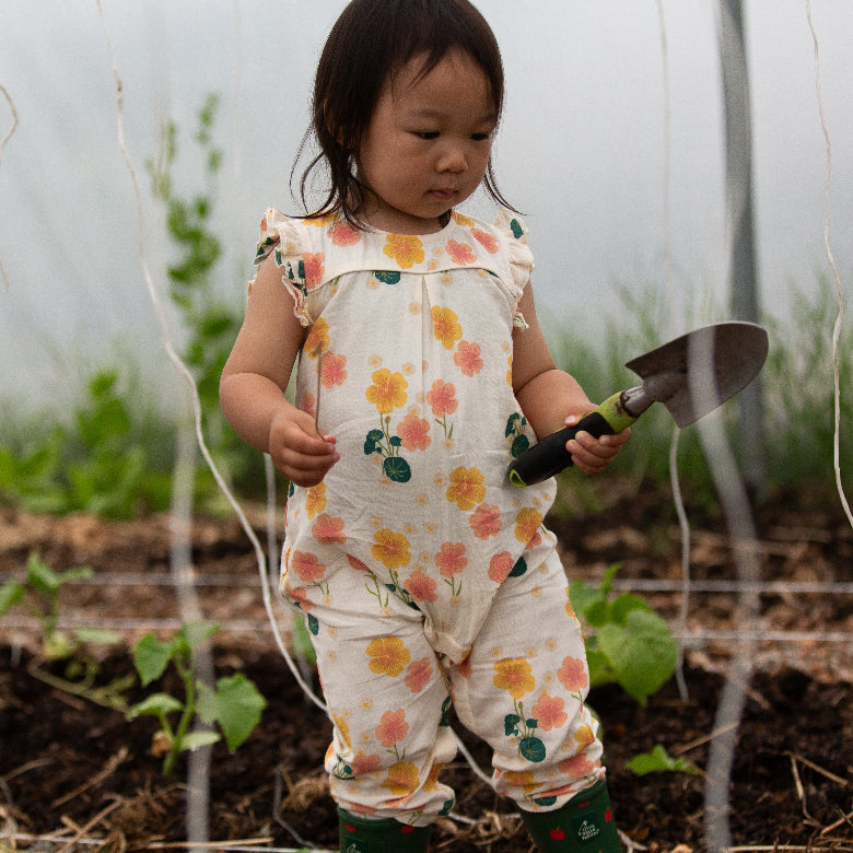 Child in a floral jumpsuit holding a small gardening tool in a garden setting