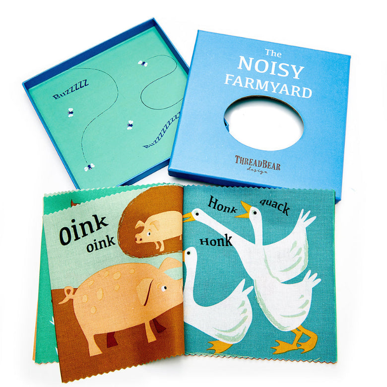 ThreadBear Designs 'The Noisy Farmyard' baby book set, showing the blue cardboard cover with a circular cutout, the teal inner board, and two colorful fabric-covered cards featuring a pig and geese.