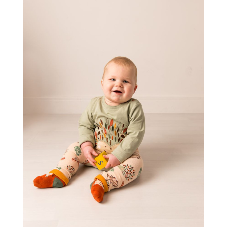 A baby sitting on a light beige studio floor wearing a sage green long-sleeve top with a colorful geometric pattern on the front, matching patterned pants, and orange socks.