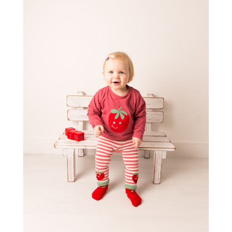 A toddler girl sitting on a white wooden bench wearing a red long-sleeve shirt with a strawberry graphic, red and white striped leggings with strawberry details, and red socks.