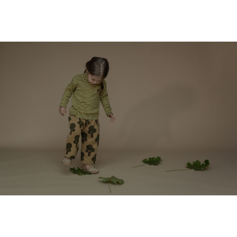 A young child wearing a green and yellow striped long-sleeve top and brown pants with a green leaf pattern, standing on a beige studio floor with a few green leaves scattered around.