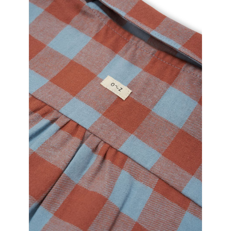 Close-up of a plaid garment fabric in muted rust orange and light blue, with a small fabric label reading '0~Z' attached near the waistband.
