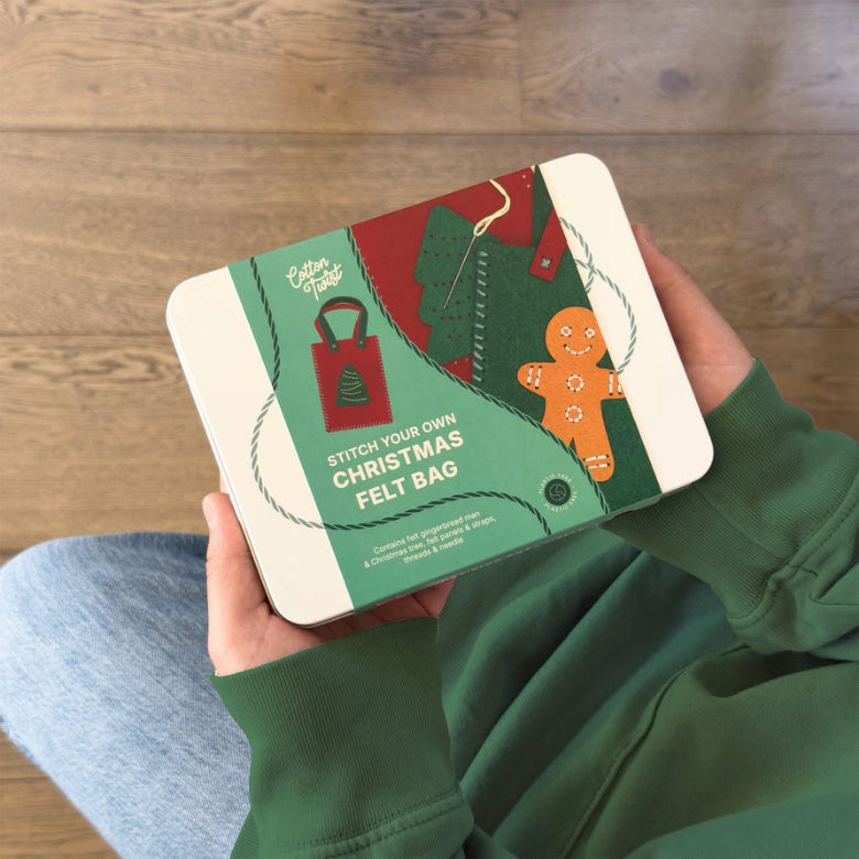 A person in a green sweatshirt holds a square metal tin featuring a Christmas-themed felt bag design. The tin has an illustrated green felt bag, red stitching details, a gingerbread man, and the text 'STITCH YOUR OWN CHRISTMAS FELT BAG.