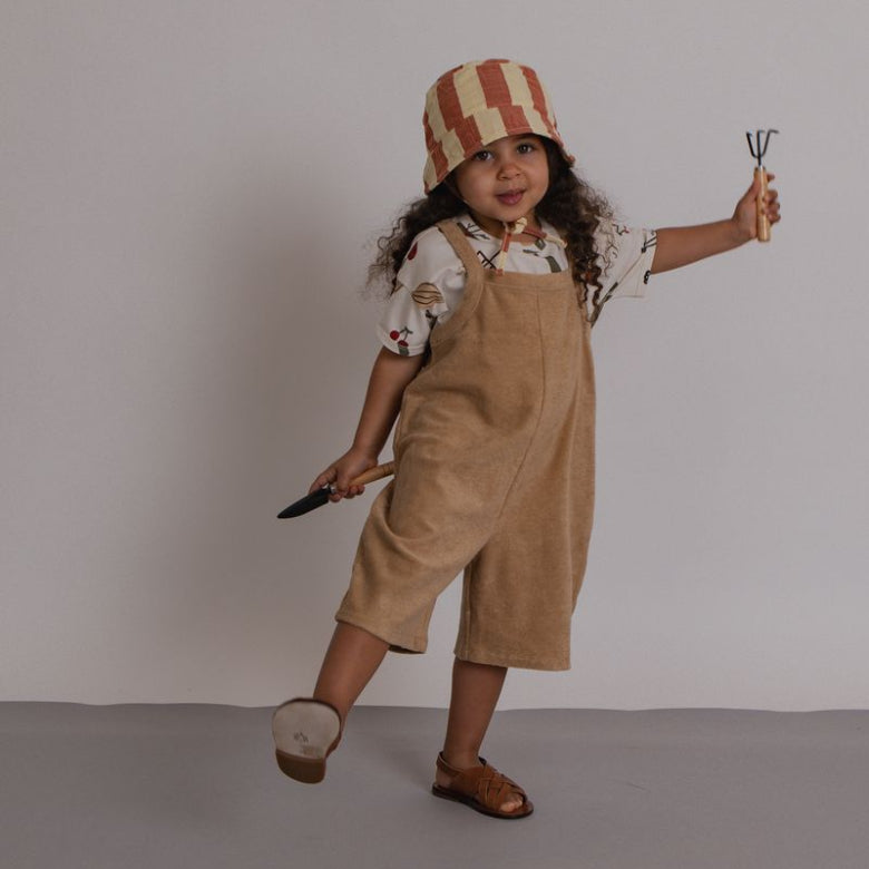 Child wearing a striped hat and beige overalls holding gardening tools against a plain background
