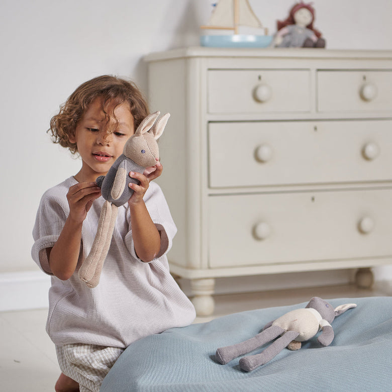 A young child sits on a blue cushion in a nursery, holding a plush gray and beige stuffed bunny. A matching stuffed bunny rests on the cushion in the foreground, with a cream-colored wooden dresser in the softly blurred background.